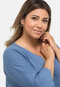 Maglione blu lavorato a maglia con un ampio scollo. Il soggetto indossa orecchini a cerchio dorati, un materiale leggermente testurizzato e maniche morbide. Tono della pelle uniforme.