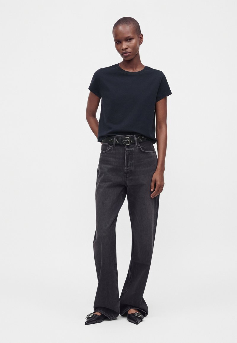 Femme aux cheveux courts portant un t-shirt noir, un jean large sombre taille haute, une ceinture noire et des chaussures noires pointues sur fond blanc.