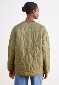 Olivno zelena prešita jakna z valovitim šivanim vzorcem, okroglim izrezom in dolgimi rokavi, kombinirana s sproščenimi modrim džins hlačami.