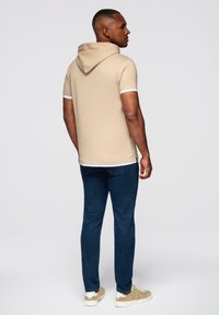 Muž stojící, mírně otočený na stranu, oblečený v béžové mikině s krátkým rukávem, modrých džínech a béžových teniskách na jednobarevném pozadí.