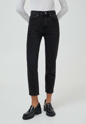Femme portant un jean noir taille haute à jambes droites, des mocassins noirs épais et un haut blanc à manches longues rentré, debout avec les mains dans le dos.