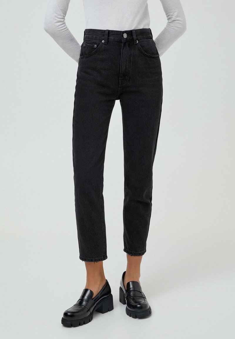 Femme portant un jean noir taille haute à jambes droites, des mocassins noirs épais et un haut blanc à manches longues rentré, debout avec les mains dans le dos.