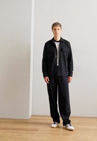Uomo in piedi su un pavimento in legno che indossa giacca e pantaloni neri, una maglietta bianca e sneakers bianche con strisce nere, di fronte a un muro bianco semplice.