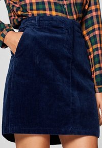 Jupe en velours côtelé bleu marine avec une taille ajustée, des poches latérales et une coupe droite jusqu'à mi-cuisse. Texture douce avec un subtil détail en côtes.