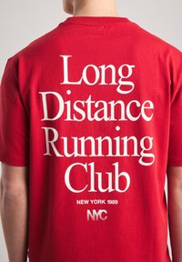 Punainen puuvillapaita, jossa on suuri valkoinen teksti: "Pitkän matkan juoksuklubi," "NEW YORK 1989," ja "NYC" raidallisin efektein.