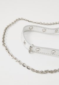 Culeră albă din piele cu accesorii din argint și gromete circulare, conectată la o lanț de argint, având finisaje netede și texturate.