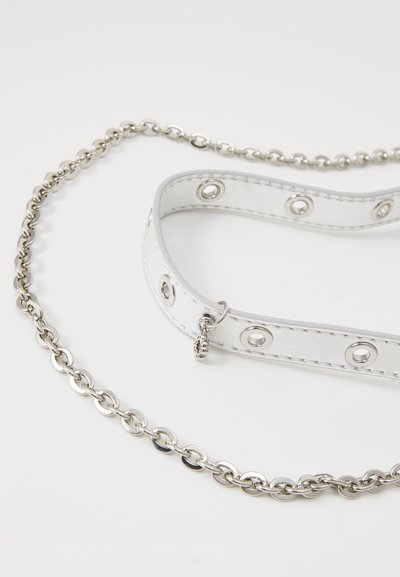 Vit läderhalsband med silverdetaljer och runda öglor, kopplat till en silverkedja, med släta och strukturerade ytor.