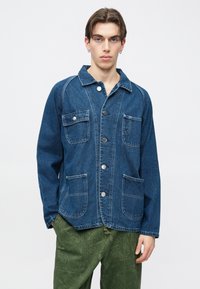 Chaqueta de mezclilla azul medio con cuello de solapa, cierre frontal con botones, dos bolsillos en el pecho y dos bolsillos laterales. Presenta costuras blancas.