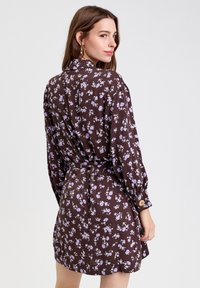 Robe florale brune à manches longues, taille cintrée et poignets boutonnés. Présente de petits motifs de fleurs violettes et blanches partout.