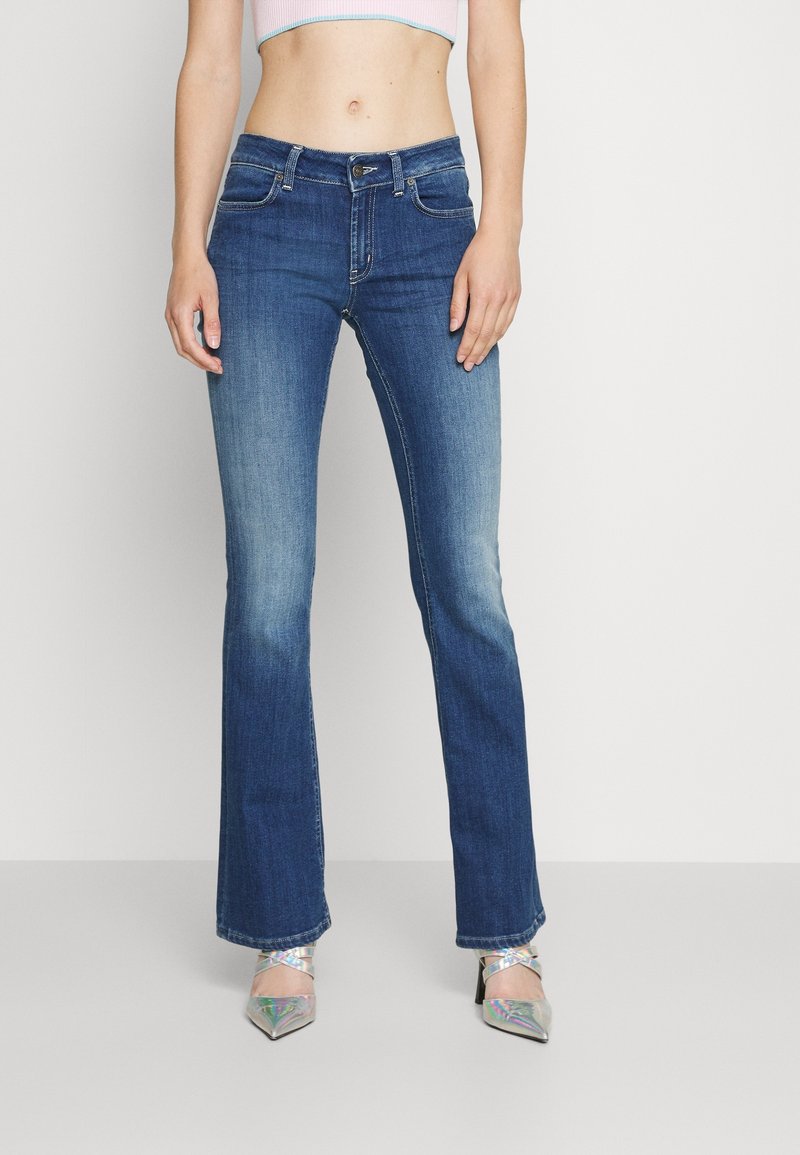 Jeans de campana en denim azul medio, con cintura estándar, cinco bolsillos y un ligero desgastado. Llevados con tacones metálicos de punta afilada.