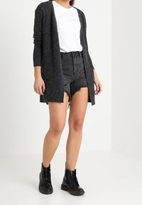 Ciemnoszary długi kardigan wykonany z fakturowanego dzianiny, noszony na białej koszulce i czarnych połatanych szortach, uzupełniony czarnymi sznurowanymi botkami.