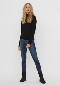Svart langermet topp, slim-fit, kombinert med blå skinny jeans. Modellen har på seg svarte snørebånds ankelstøvler. Enkel design mot en nøytral bakgrunn.