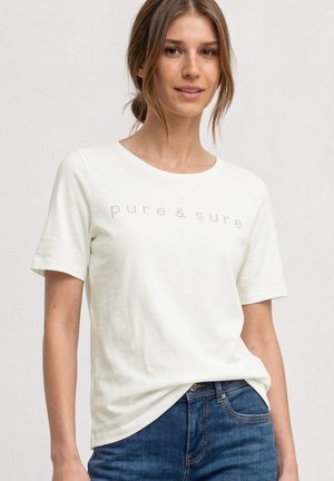 Ung kvinde med lystbrunt hår iført en hvid t-shirt med teksten "pure & sure" og blå jeans, stående mod en ensfarvet baggrund.