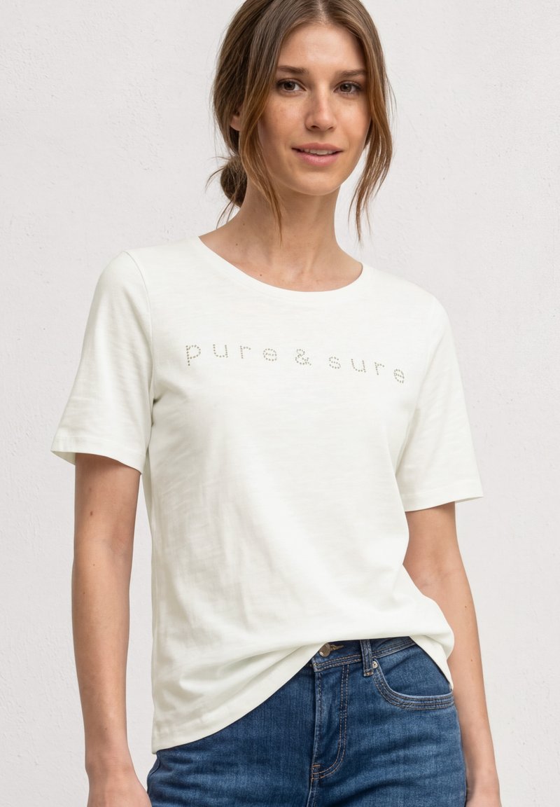 Jeune femme aux cheveux châtain clair portant un t-shirt blanc avec le texte « pure & sure » et un jean bleu, debout devant un fond uni.