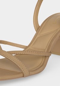 Bézs színű, sarokkal ellátott szandál vékony, varrott pántokkal és párnázott talpbéléssel, oldalnézetből a cipő eleje és sarka látható.