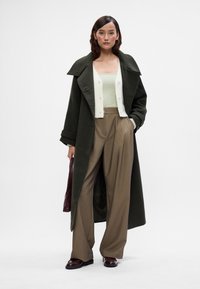 Mulher vestindo um casaco longo verde escuro, um colete creme, uma blusa verde clara, calças largas castanhas e sapatos burgundy, segurando uma bolsa combinando.