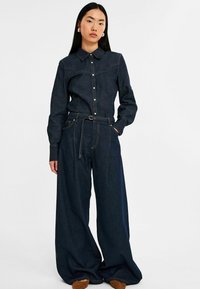 Mørk denim outfit bestående af en skjorte med knapper og krave samt bukser med brede ben, accentueret med kontrasterende syninger og et bælte om taljen.