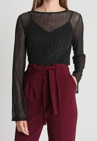 Top lungo a maniche trasparenti nere con strisce verticali, indossato sopra un sottogono nero. Abbinato a pantaloni bordeaux a vita alta con una cintura legata.