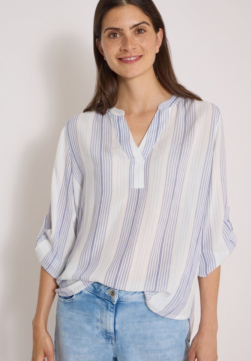 Femme aux longs cheveux bruns portant un chemisier blanc à rayures verticales bleues et un jean bleu clair, souriant devant un fond uni.