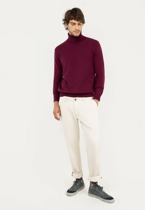 Hombre con un jersey de cuello alto burdeos, pantalones blancos remangados y zapatillas deportivas gris oscuro, de pie frente a un fondo claro y liso.