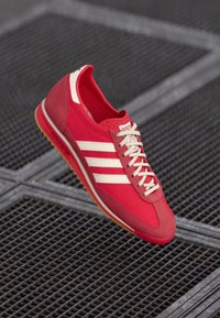 Zapatilla deportiva roja con rayas blancas, parte superior de tela, suela de goma y puntera redondeada. Cierre con cordones y acentos sintéticos.