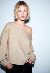 Beżowy wełniany sweter o kroju one-shoulder, z prążkowanymi mankietami i luźnym krojem, zestawiony z ciemnoniebieskimi dżinsami. Czyste, minimalistyczne tło.