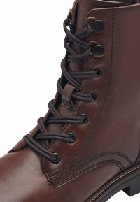 Botte en cuir marron avec une texture lisse, dotée d'œillets en métal, de lacets marron foncé et d'une semelle contrastante. Couture détaillée visible.