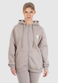 Lichtgrijze zip-up hoodie gemaakt van zachte stof, met een kangoeroezak, verstelbare capuchon en een geborduurd logo op de borst.