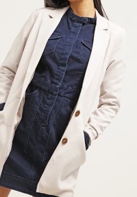 Kermanvalkoinen bleiseri, jossa on räätälöity istuvuus, yhdistettynä tummansiniseen farkkumekkoon, jossa on korkea kaulus, napitus ja päälle ommellut taskut.