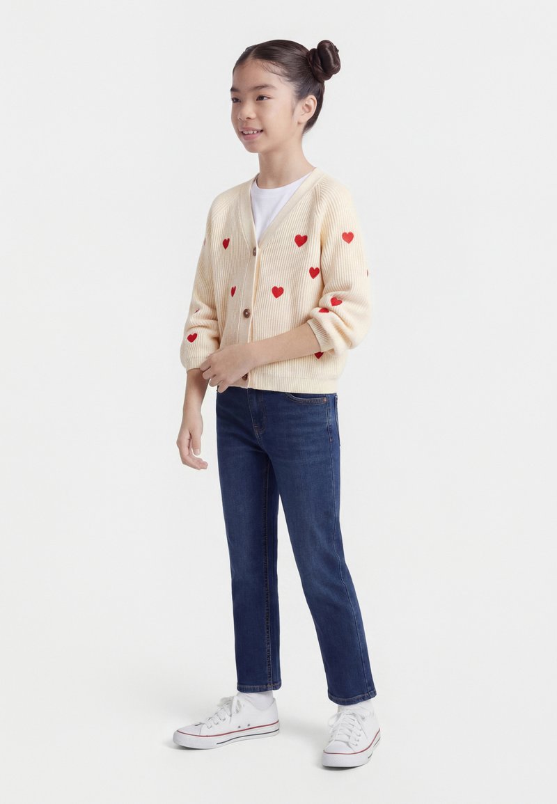 Νεαρή κοπέλα που στέκεται, φοράει κρεμ ζακέτα με κόκκινη καρδιές, λευκό πουκάμισο, μπλε τζιν και λευκά αθλητικά παπούτσια, χαμογελώντας ελαφρώς προς τα πλάγια.