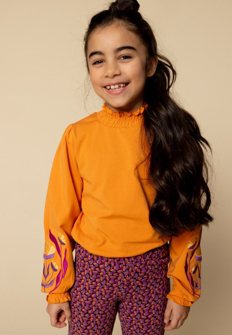 Jong meisje met lang donker haar in een oranje top met bloemenborduursel op de mouwen en paarse gestreepte broek, lachend tegen een beige achtergrond.