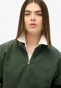 Sudadera de cremallera verde oscuro con un cuello blanco. Presenta una cremallera frontal y un logo bordado en el pecho. Textura de tela suave.
