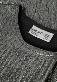 Szary teksturowany top o prążkowanym wzorze z metalicznymi ozdobami. Dekolt jest miękki, z czarną wewnętrzną lamówką oraz metką z informacjami o pielęgnacji.