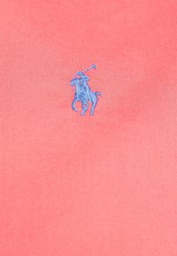 Pikétröja i korallfärgad tyg med en slät yta, prydd med en blå broderad logotyp av en häst och ryttare.