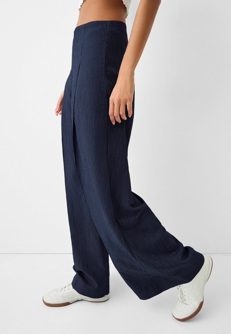 Pantaloni a gamba larga blu realizzati in tessuto a coste con righe verticali, dotati di vita alta e tasche laterali, abbinati a sneakers bianche.