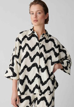 Jeune personne aux cheveux courts portant une tenue ample couleur crème avec des motifs en zigzag noirs audacieux, debout devant un fond clair uni.