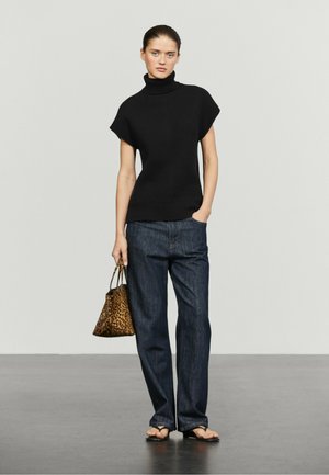 Frau trägt schwarzen ärmellosen Rollkragen, dunkelblaue Jeans, hält eine Handtasche mit Leopardenmuster und trägt schwarze Sandalen auf grauem Boden.