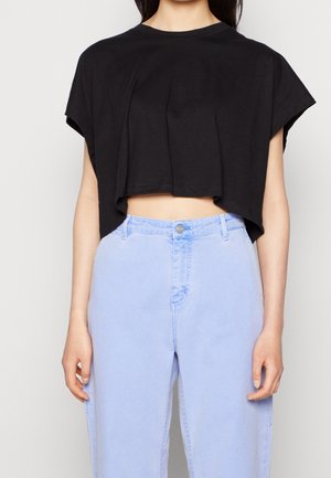 Nainen, jolla on yllään löysä musta lyhyt toppi ja korkeavyötäröiset vaaleansiniset farkut, seisoo vaaleaa taustaa vasten.