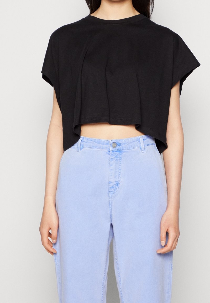 Femme portant un haut court noir ample et un jean taille haute bleu clair, se tenant contre un fond clair uni.