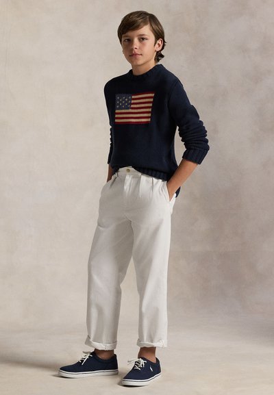 Ragazzo in piedi con le mani in tasca, indossa un maglione blu navy con la bandiera americana, pantaloni bianchi e scarpe da ginnastica blu navy su sfondo neutro.