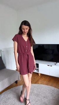 Γυναίκα με βυσσινί ολόσωμη φόρμα με κουμπιά και σανδάλια που στέκεται σε στρογγυλό γκρι χαλί σε σαλόνι με τηλεόραση και καναπέ.