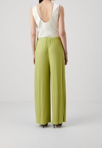 Hellgrüne, plissierte Hosen mit einem elastischen Bund, kombiniert mit einem weißen, tief ausgeschnittenen Tanktop. Das Modell trägt marineblaue Absätze. Einfacher Hintergrund.
