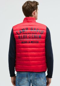 Rote Steppweste mit vertikalen Rauten. Auf der Rückseite befindet sich in Schwarz ein Logo. Kombiniert mit einem marineblauen Langarmshirt und Jeans.
