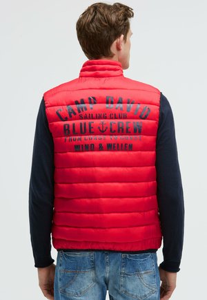Gilet imbottito rosso con sezioni trapuntate verticali. Presenta un testo con logo nero sul retro. Abbinato a una maglietta a maniche lunghe blu navy e jeans di denim.