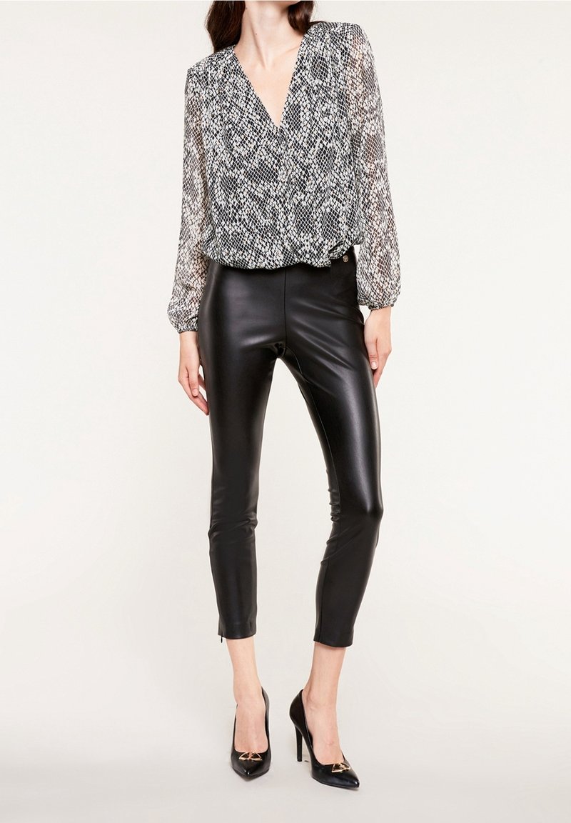 Femme portant un chemisier à manches longues à motifs noir et blanc, un pantalon en cuir noir et des chaussures à talons noirs sur un fond uni.