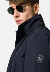 Granatowa kurtka z wysokim kołnierzem, szczegółowymi guzikami i okrągłą naszywką z logo na rękawie. Model nosi okrągłe okulary przeciwsłoneczne typu aviator.