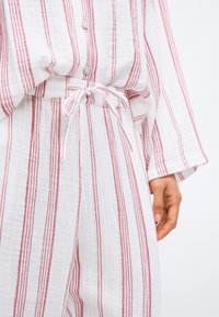 Białe bawełniane ubranie w czerwone pionowe paski; zawiera zapięcie na guziki u góry z luźnym krojem oraz wiązany pas w spodniach, podkreślając miękki materiał.