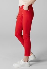 Jean skinny rouge en tissu mélangé coton, avec un design classique à cinq poches et des accessoires en métal argenté, associés à des baskets blanches.