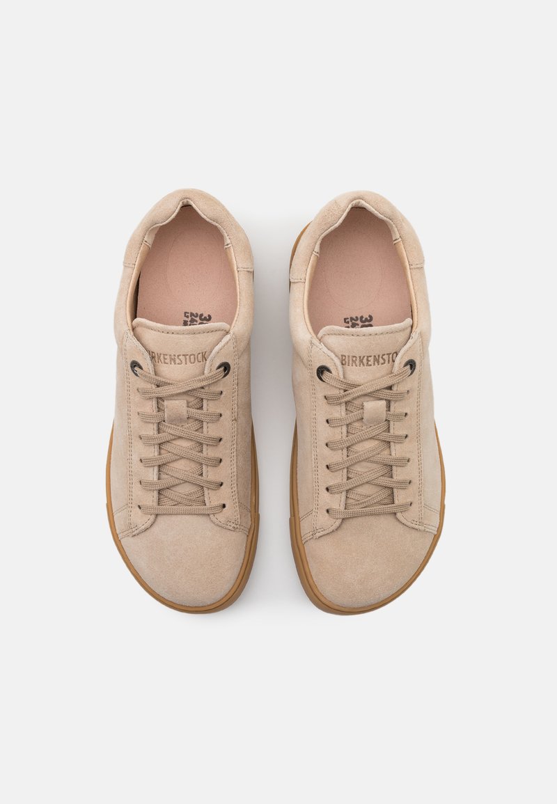 Beige mockasinsneakers med snörning, texturerad yta och gummisula. Varumärke på tungan och minimala sömnadsdetaljer.