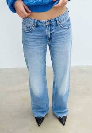 Personne portant un jean large bleu clair et des chaussures noires à bout pointu, remontant un pull en tricot bleu, debout sur un sol en béton.